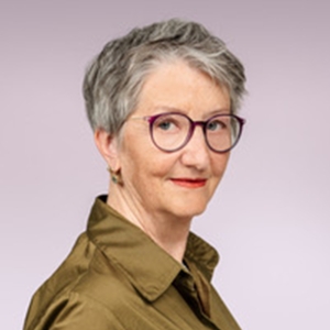 Das Gesicht einer Frau mit kurzen, grauen Haaren und violetter Brille und olivgrüner Bluse.
